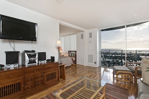 A modern living room with a flat-screen TV, wooden furniture, and a large window offering a city view.