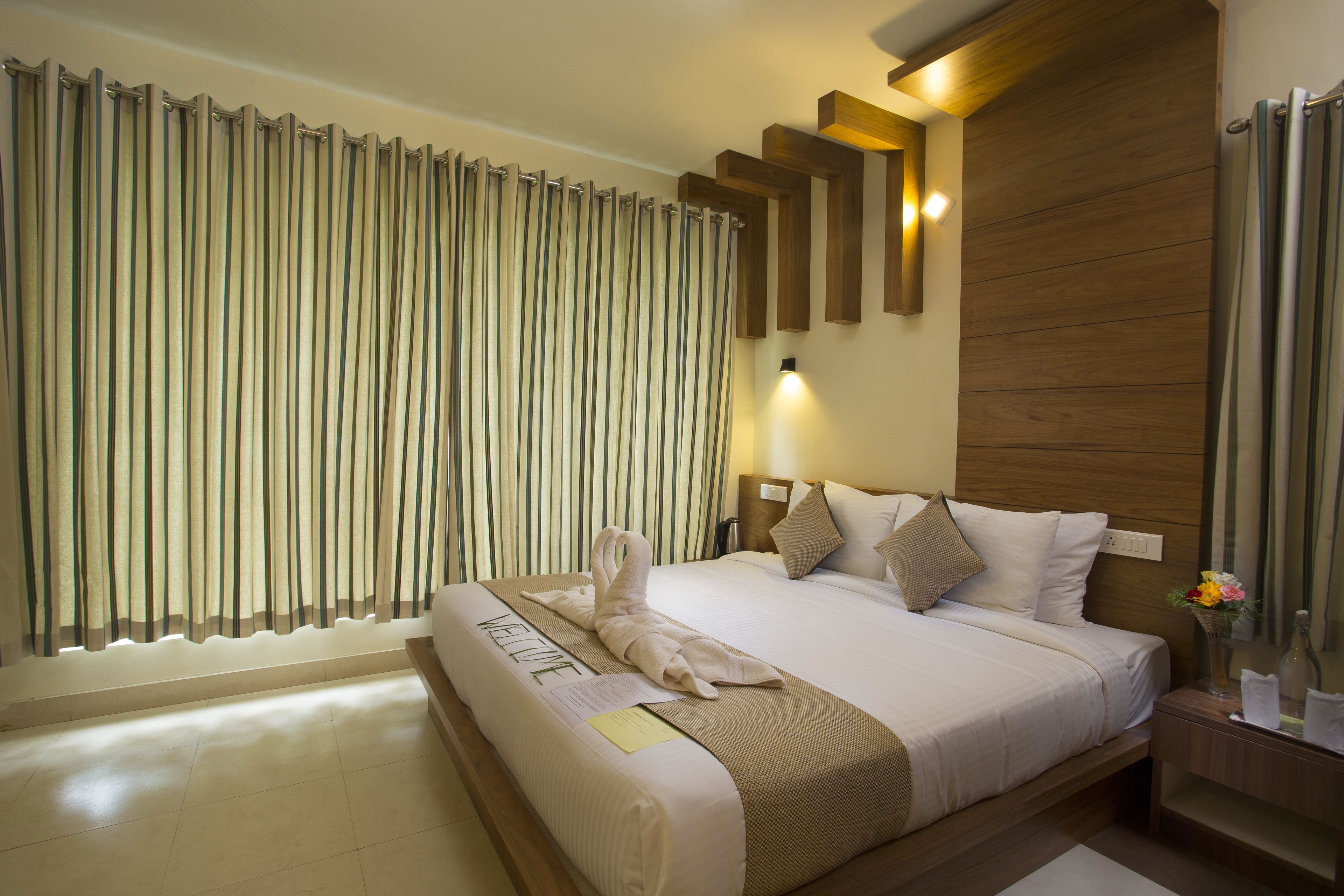 A hotel room with a large bed, a wooden headboard, a nightstand with a vase of flowers, and a window with curtains.