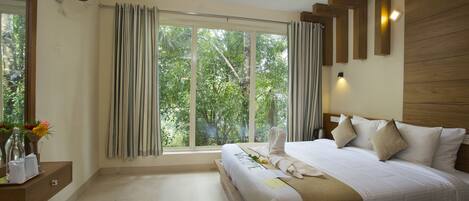 Quarto de hotel com uma cama grande, uma mesinha de cabeceira com um vaso de flores, uma janela com cortinas e um ventilador fixado na parede.