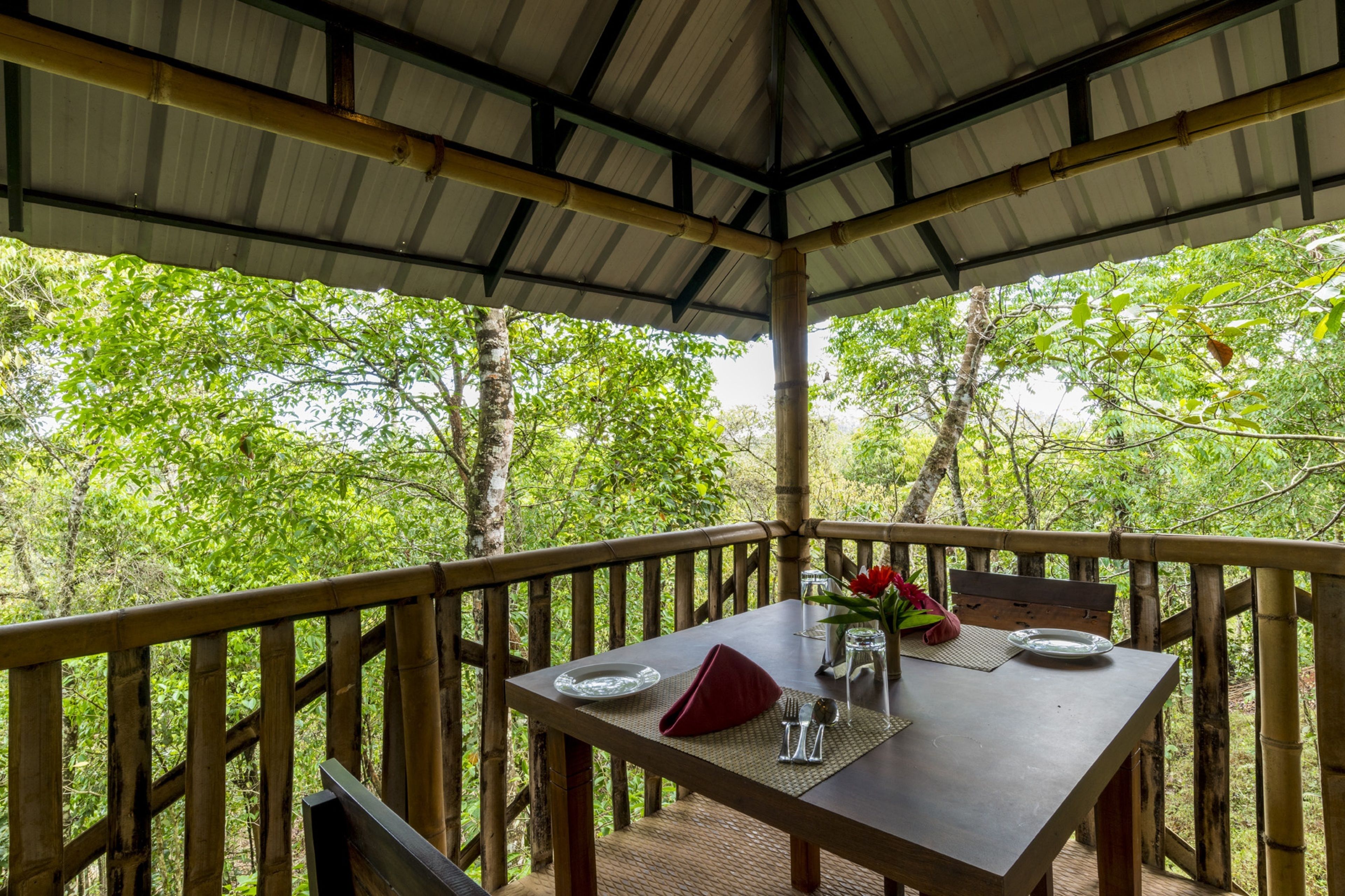An outdoor dining area with a table set for two, surrounded by a wooden deck and bamboo railings, overlooking a lush forest.
