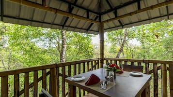 An outdoor dining area with a table set for two, surrounded by a wooden deck and bamboo railings, overlooking a lush forest.