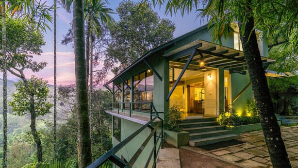 Un bâtiment moderne doté d’une entrée en verre, entouré d’une végétation luxuriante et adossé à un paysage montagneux.