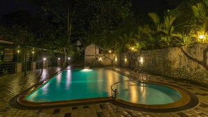 Une piscine bordée de pierres, entourée d’un mur de pierres orné de motifs décoratifs, et une petite maison en arrière-plan.