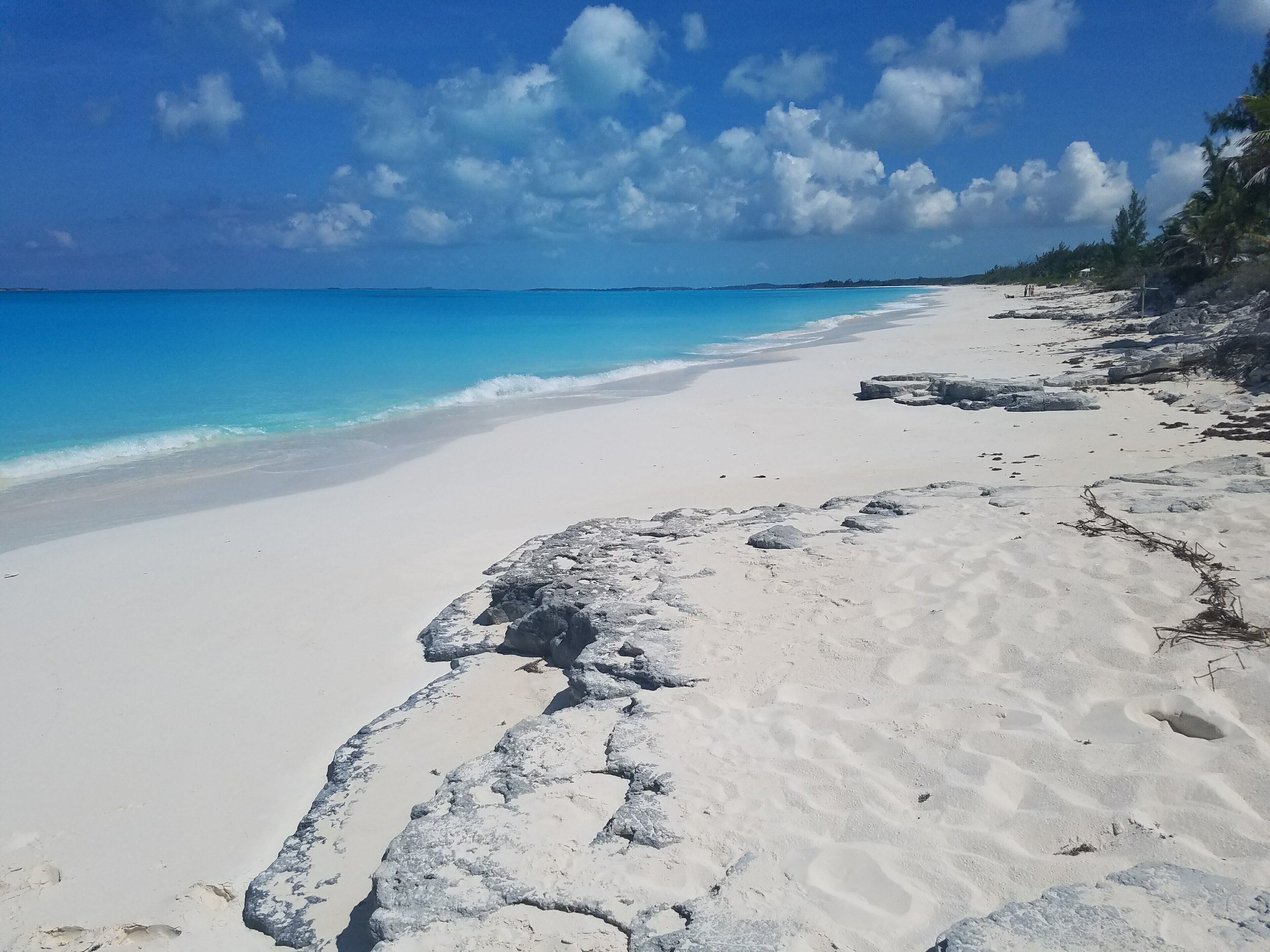 Een strand met wit zand en grote rotsen, helderblauw water en enkele bomen.