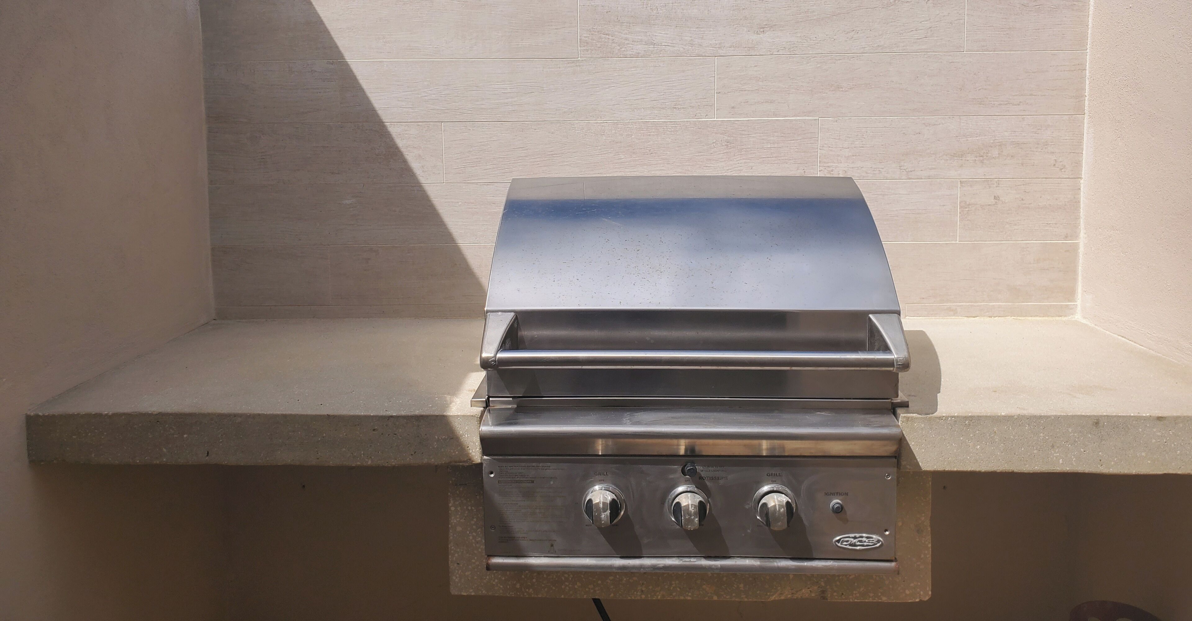 A stainless steel grill with three control knobs and a digital display, mounted on a concrete countertop against a tiled wall.