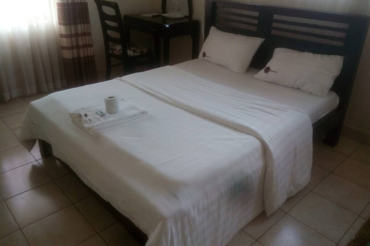 Una cama doble bien hecha con sábanas blancas, una mesita con una silla y una ventana con cortinas.