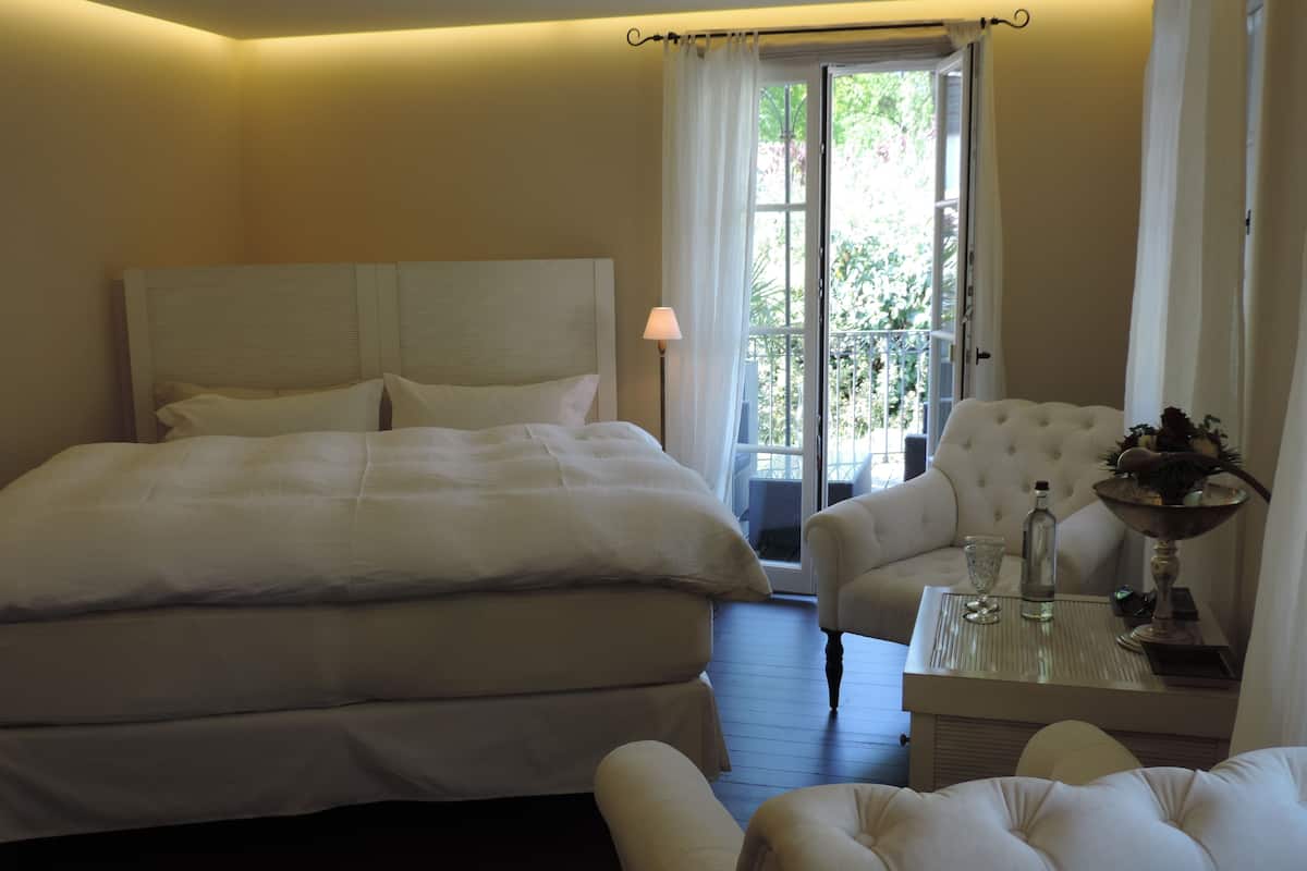 A hotel room with a large bed, a sitting area with a chair and a table, and a balcony with a view of greenery.