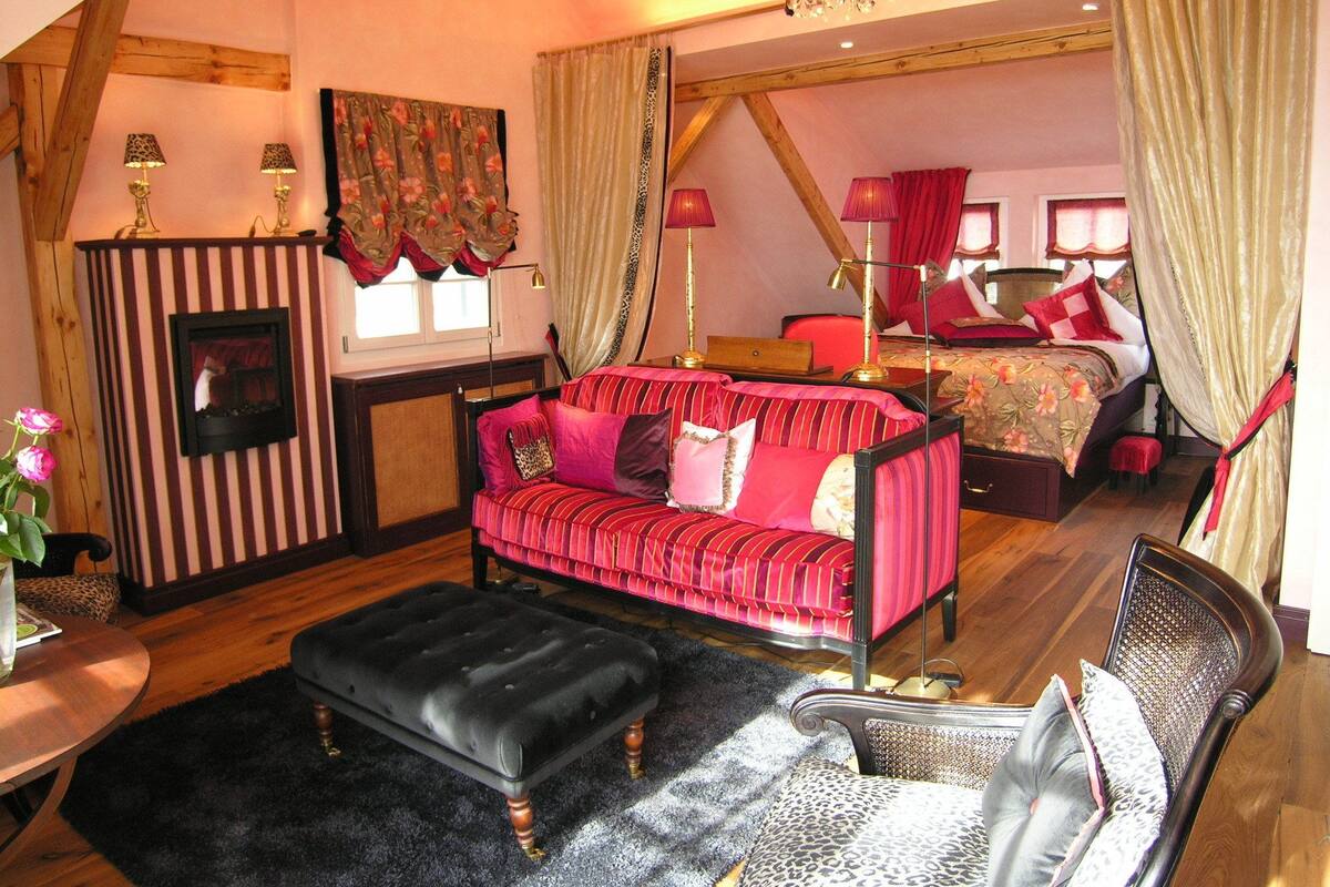 A cozy bedroom with a large bed, a sitting area with a sofa and ottoman, a fireplace, and a view of the bedroom through a window.