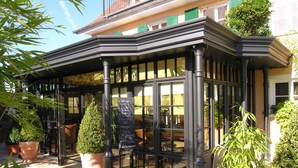 A corner building with a black awning, large windows, and potted plants. - Pension Am Bodensee (Kressbronn)