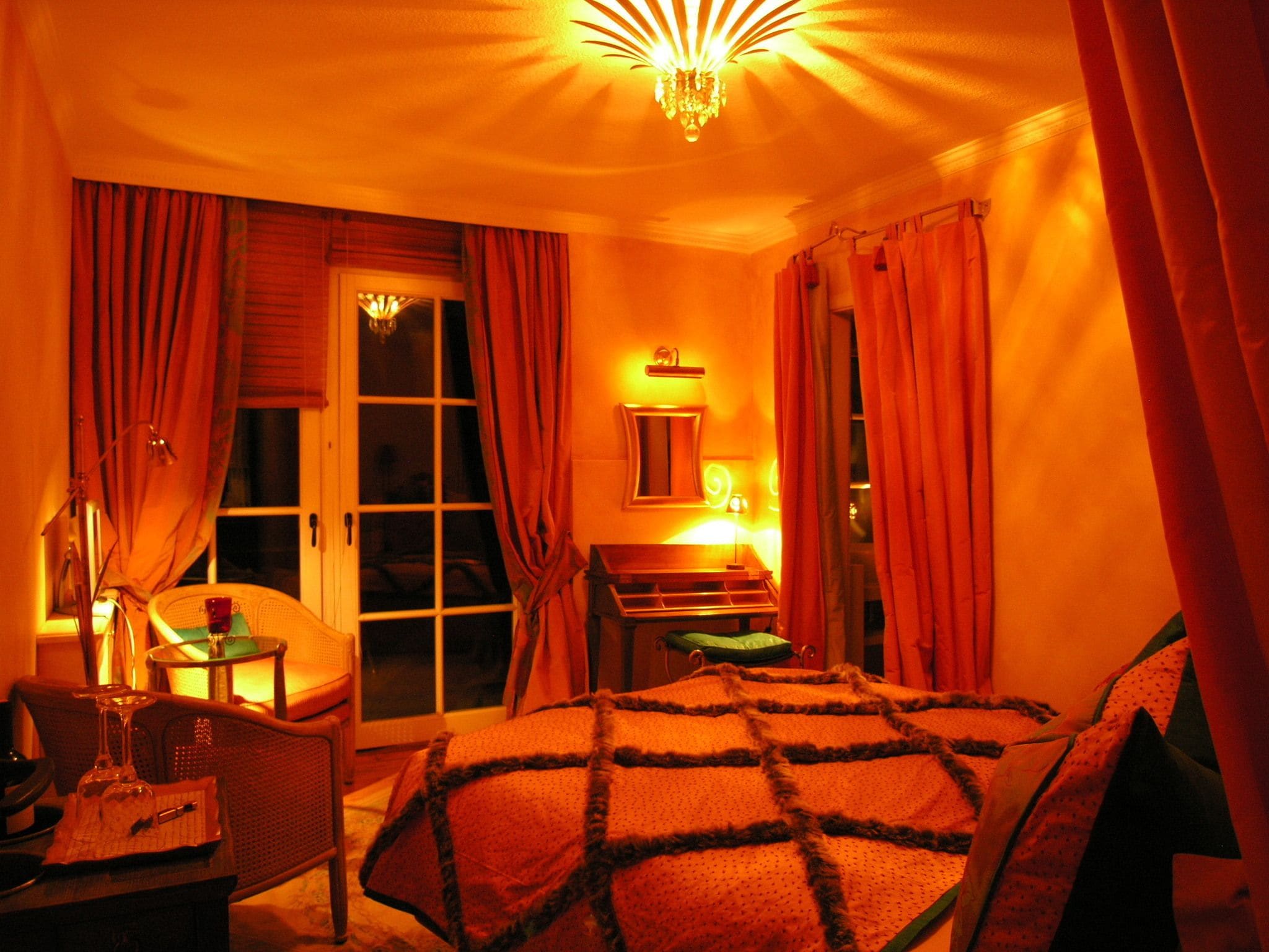 A hotel room with a bed, a desk, a chair, a window with curtains, and a chandelier.