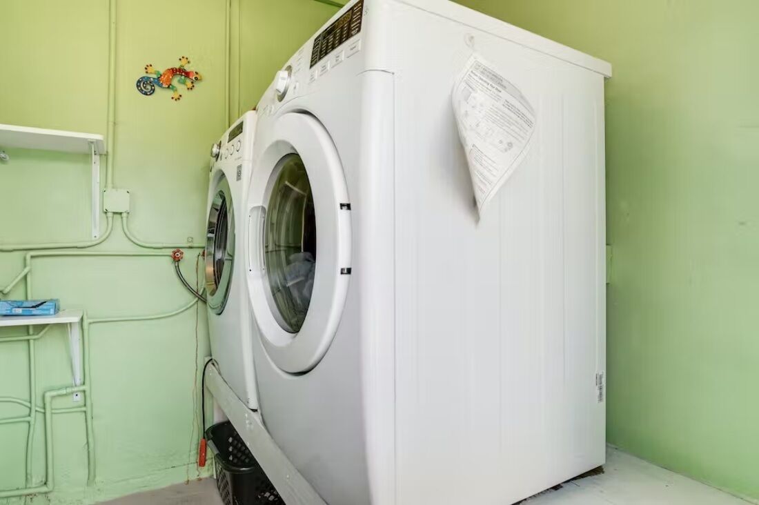 Una lavadora blanca de carga frontal, compacta, en una habitación con paredes verde claro y un pequeño estante.