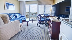 Habitación de hotel con sofá, una mesa de comedor con sillas, televisión y vistas al mar a través de amplios ventanales.