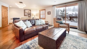 Ein gemütliches Wohnzimmer mit einer Ledersofa, einem Holztisch und Blick auf eine verschneite Landschaft durch große Fenster.