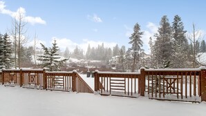 Eine Holzterrasse mit schneebedeckten Geländern, die zu einer verschneiten Landschaft mit Bäumen und einem Gebäude im Hintergrund führt.