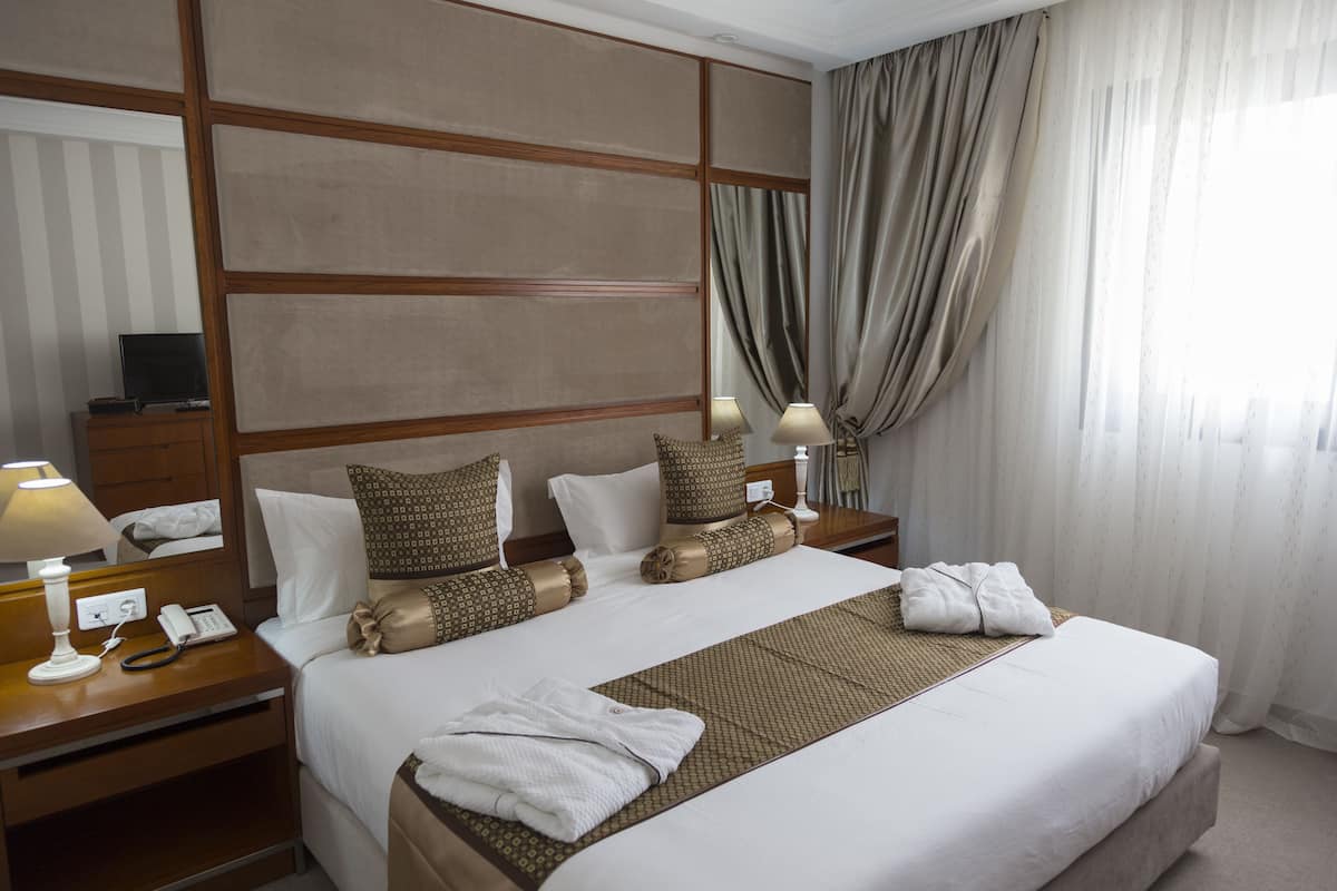 A hotel room with a large bed, two bedside tables, a lamp, a telephone, and a window with curtains.