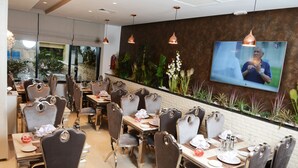A restaurant with multiple dining tables and chairs, a television mounted on the wall, and a variety of plants along the wall. - Marigold Hotel (Tunis)
