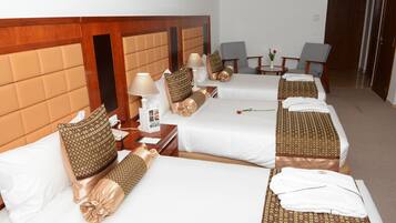 A hotel room with three beds, each with a headboard, bedside tables, lamps, and pillows. There are two chairs and a small table with a vase of flowers.