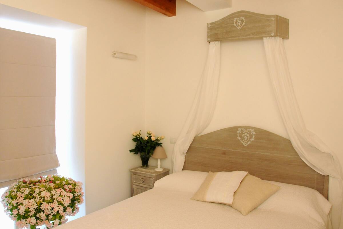 A bedroom with a wooden headboard, a canopy bed, a bedside table with a lamp and flowers, and a window with curtains.