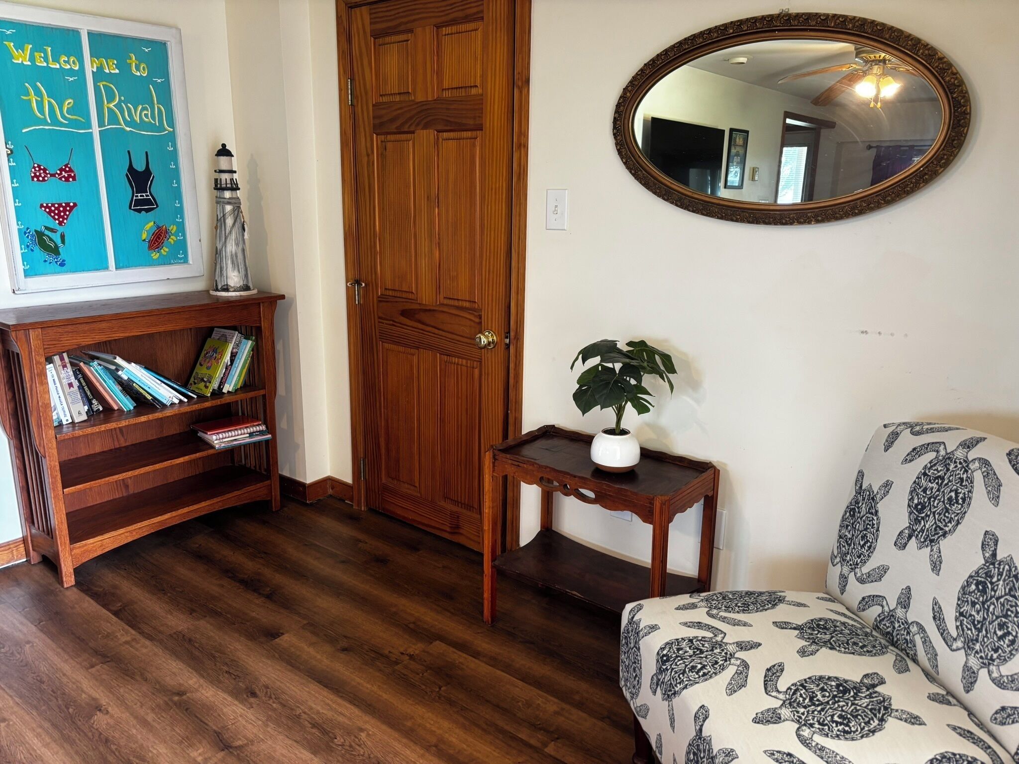 Une porte en bois, une bibliothèque remplie de livres, une petite table avec une plante, un canapé à motifs et un miroir accroché au mur.