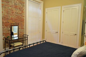 A bedroom with a brick wall, a bed with a dark cover, a window with blinds, and two white doors.