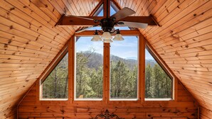 Eine Holzhütte mit geneigtem Dach, Deckenventilator und einem großen Fenster mit Blick auf einen bewaldeten Berg.