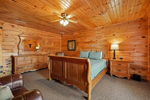 Un dormitorio con paneles de madera, una cama, un tocador, una mesita de noche, un espejo y un ventilador de techo.