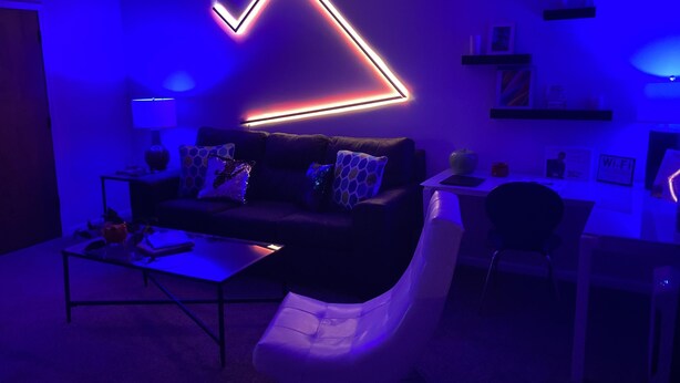 A modern living room with a dark-colored sofa, a white armchair, and a coffee table. There is a neon arrow light on the wall, a lamp on a side table, and a small shelf with items.