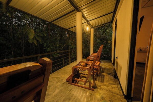 A covered outdoor seating area with wooden chairs and a bench, surrounded by lush greenery.