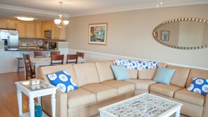 A living room with a beige sectional sofa, a white coffee table, and a dining area with wooden chairs. - Brass Rail 3 Bed, 2.5 Bath Ocean View Top Floor Closest To Beach (Tybee Island)