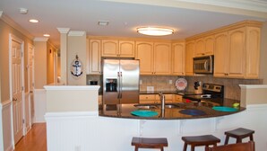 A kitchen with wooden cabinets, a stainless steel refrigerator, a microwave, and a sink. There are two wooden stools and a blue placemat on the counter. - Brass Rail 3 Bed, 2.5 Bath Ocean View Top Floor Closest To Beach (Tybee Island)