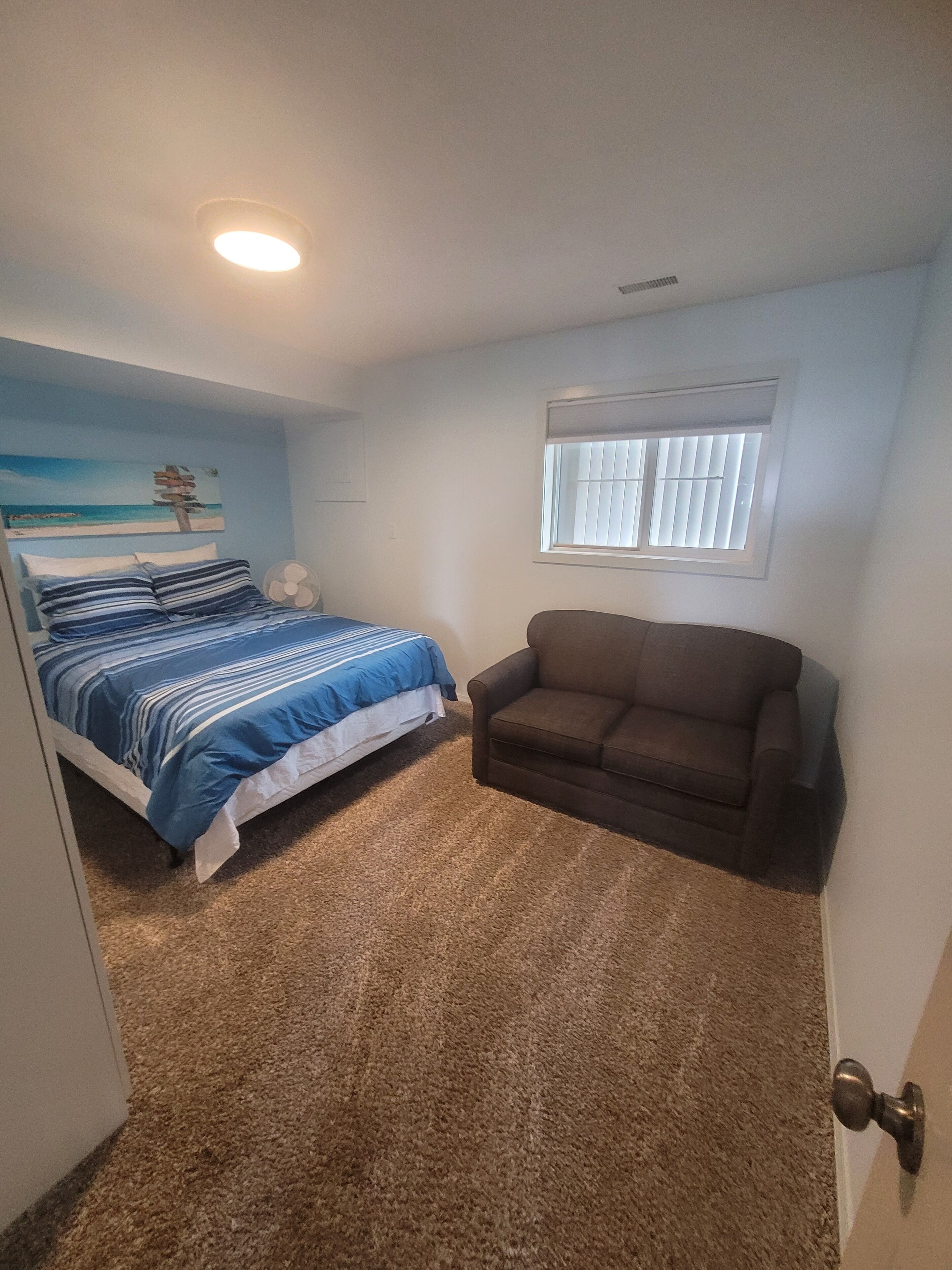 A bedroom with a bed, a brown couch, a window with blinds, and a ceiling fan.