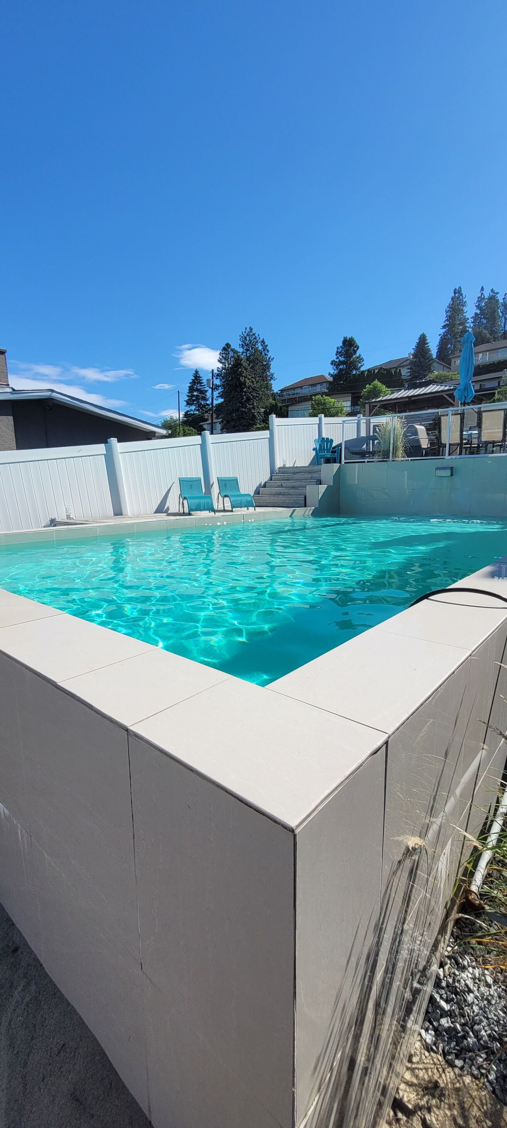 A swimming pool with clear blue water, surrounded by a white fence, and a few green chairs.