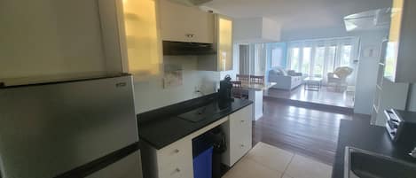 A modern kitchen with a refrigerator, stove, and sink. There is a dining area with a table and chairs. The room has large windows and a view of the outdoors.