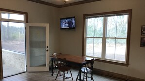 Una habitación con una mesa de comedor y sillas de madera, un televisor montado en la pared y ventanas grandes con vistas a árboles.