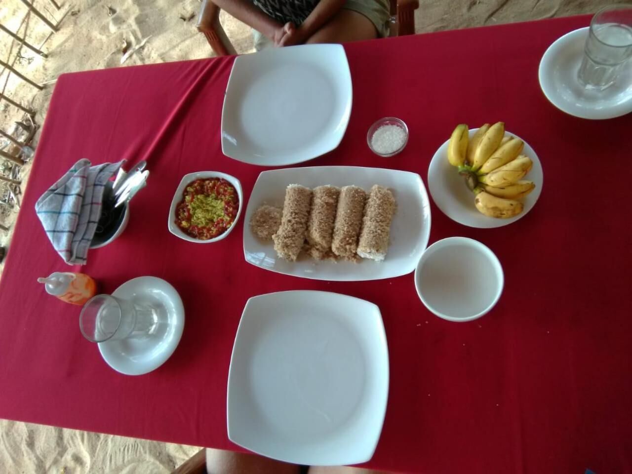 A table set with white plates, a plate of banana slices, a plate of bread rolls, a bowl of salsa, a glass of water, and a teapot.
