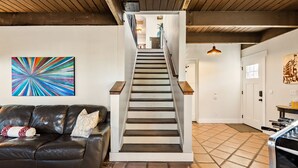 Eine Treppe führt zu einer zweiten Etage, ein schwarzes Ledersofa mit gemusterten Kissen und ein farbenfrohes abstraktes Gemälde an der Wand.