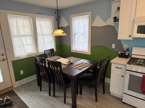 Un coin repas avec une table dressée pour quatre personnes, un luminaire suspendu et une cuisine équipée d’une cuisinière et de placards.