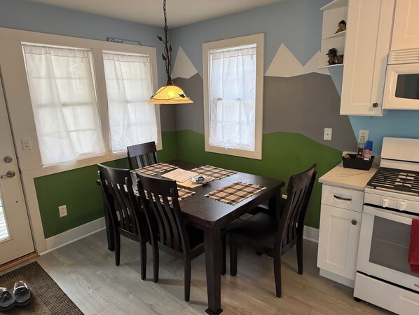 Un comedor con una mesa dispuesta para cuatro, una lámpara colgante y una cocina con estufa y alacenas.