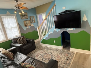 Un salon comprenant un canapé, un tapis à motifs, un téléviseur à écran plat fixé au mur et un escalier dont le mur est décoré sur le thème de la montagne.