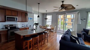 A modern kitchen with wooden cabinets, a granite countertop, and a bar area with stools. There is a dining area with a table and chairs, and a living room with a sofa and a ceiling fan.