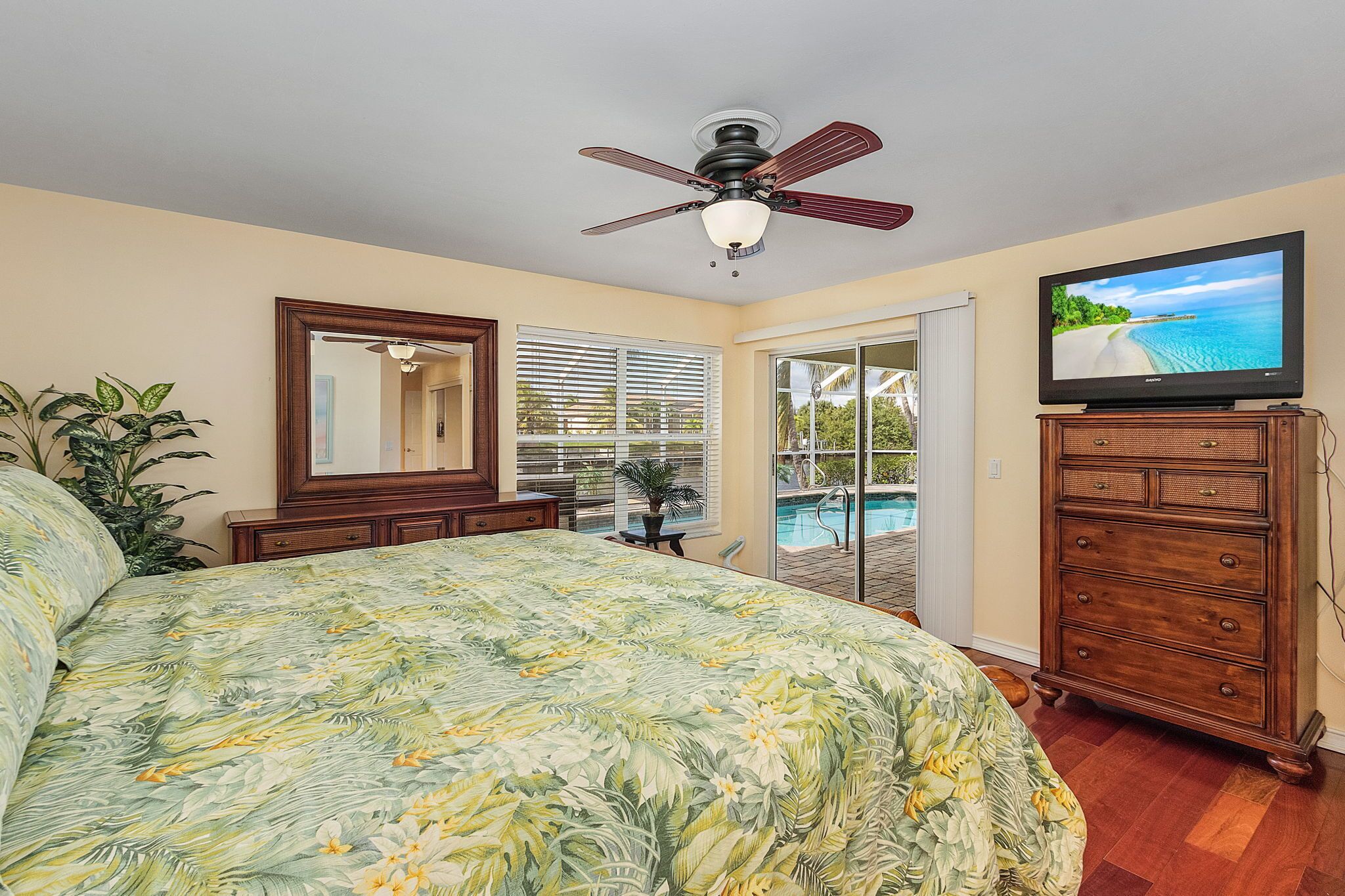 A bedroom with a bed, a ceiling fan, a television, a dresser, and a view of a pool through the door.