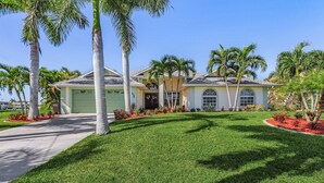 A two-story house with a green lawn, palm trees, and a garage. - Fun for the Whole Family! Pool Table, Heated Pool, Fishing Dock -Villa Serendipity-Roelens Vacations (Cape Coral)