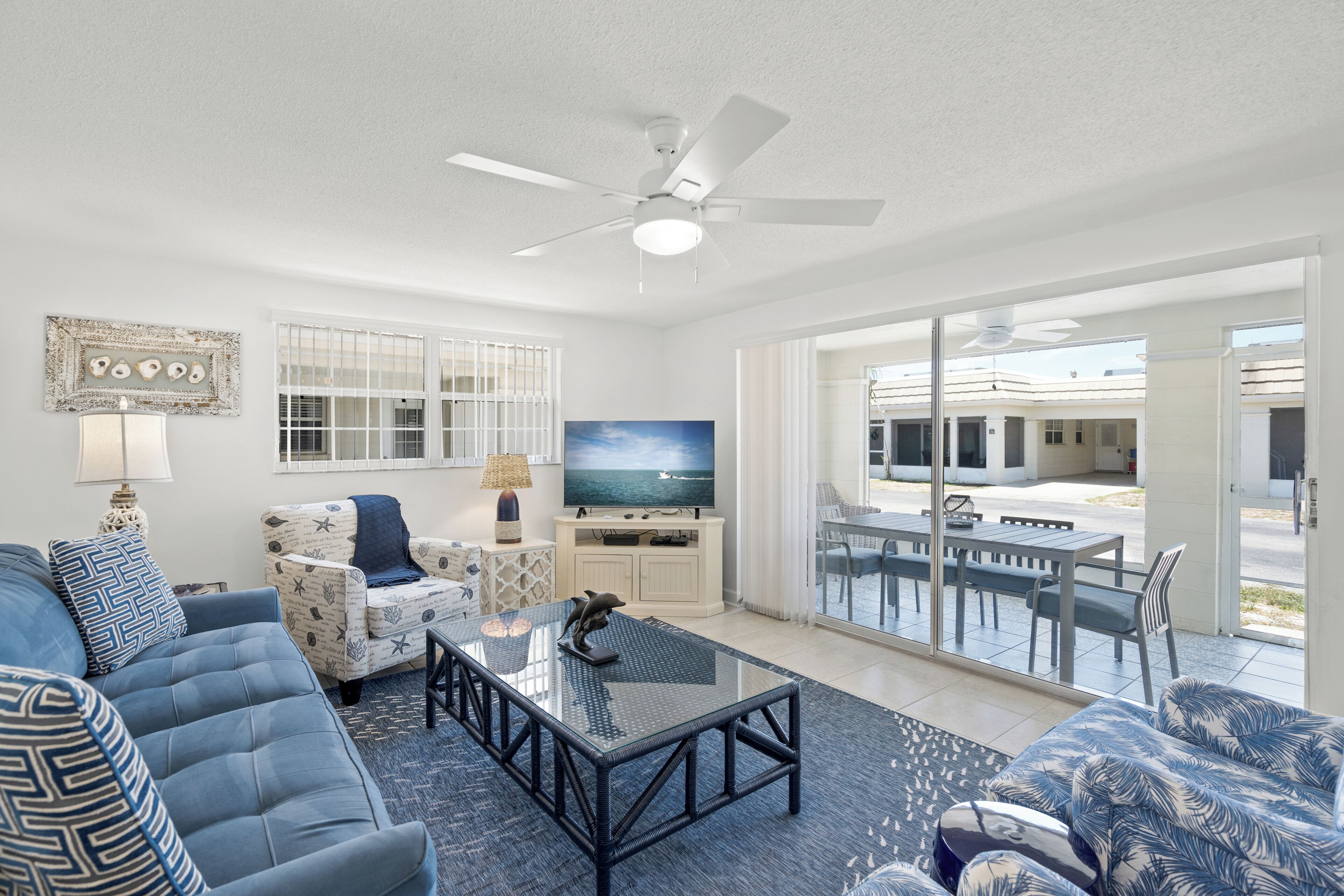A living room with a sofa, armchair, and coffee table. There is a television and a ceiling fan. The room has large windows and a view of the outside.