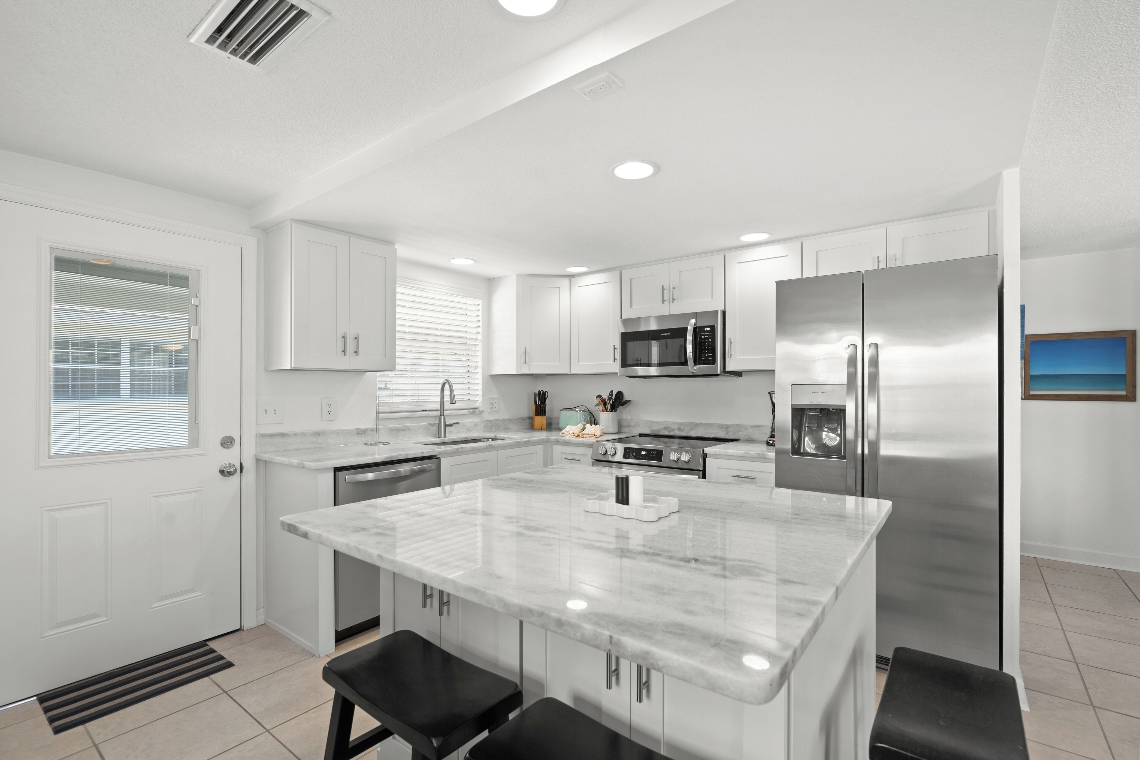 A modern kitchen with white cabinetry, a central island, stainless steel appliances, and a window with blinds.