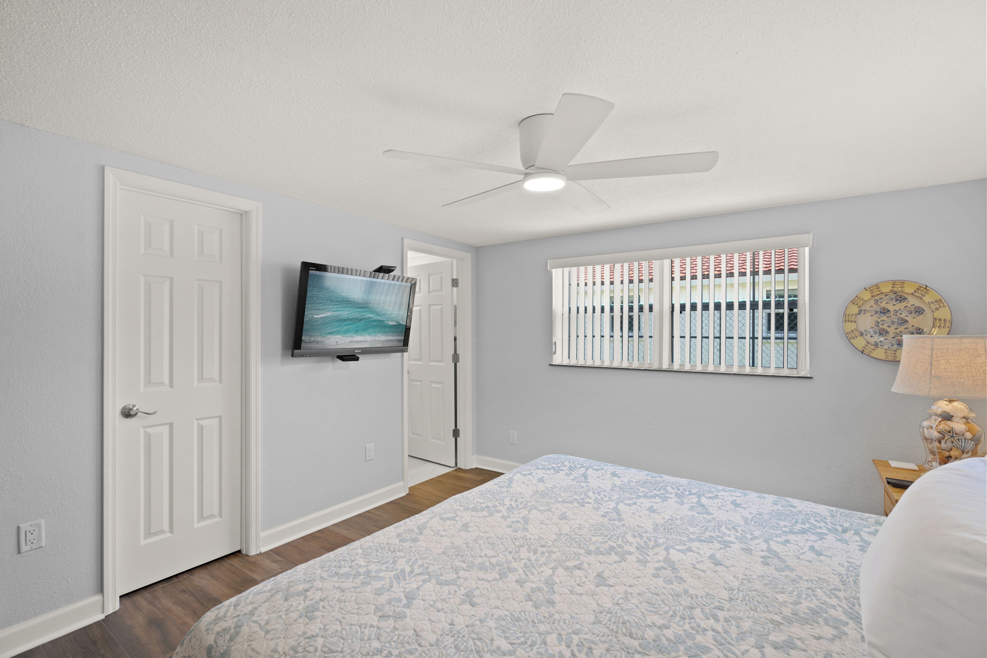 A bedroom with a bed, a ceiling fan, a TV mounted on the wall, and a window with blinds.
