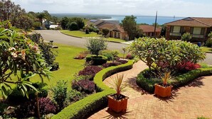 A landscaped garden with a paved pathway, various plants, and a view of the sea and houses in the background. - Best views of Jervis Bay, A/C,heating, free Internet & beautiful large apartment (Vincentia)
