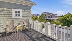 Una terraza de madera con sillas Adirondack y una mesita, baranda blanca y vistas a casas vecinas.