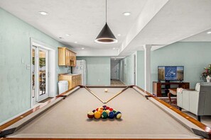 A pool table with balls, a television, and a sofa in a room with light green walls and wooden cabinets.