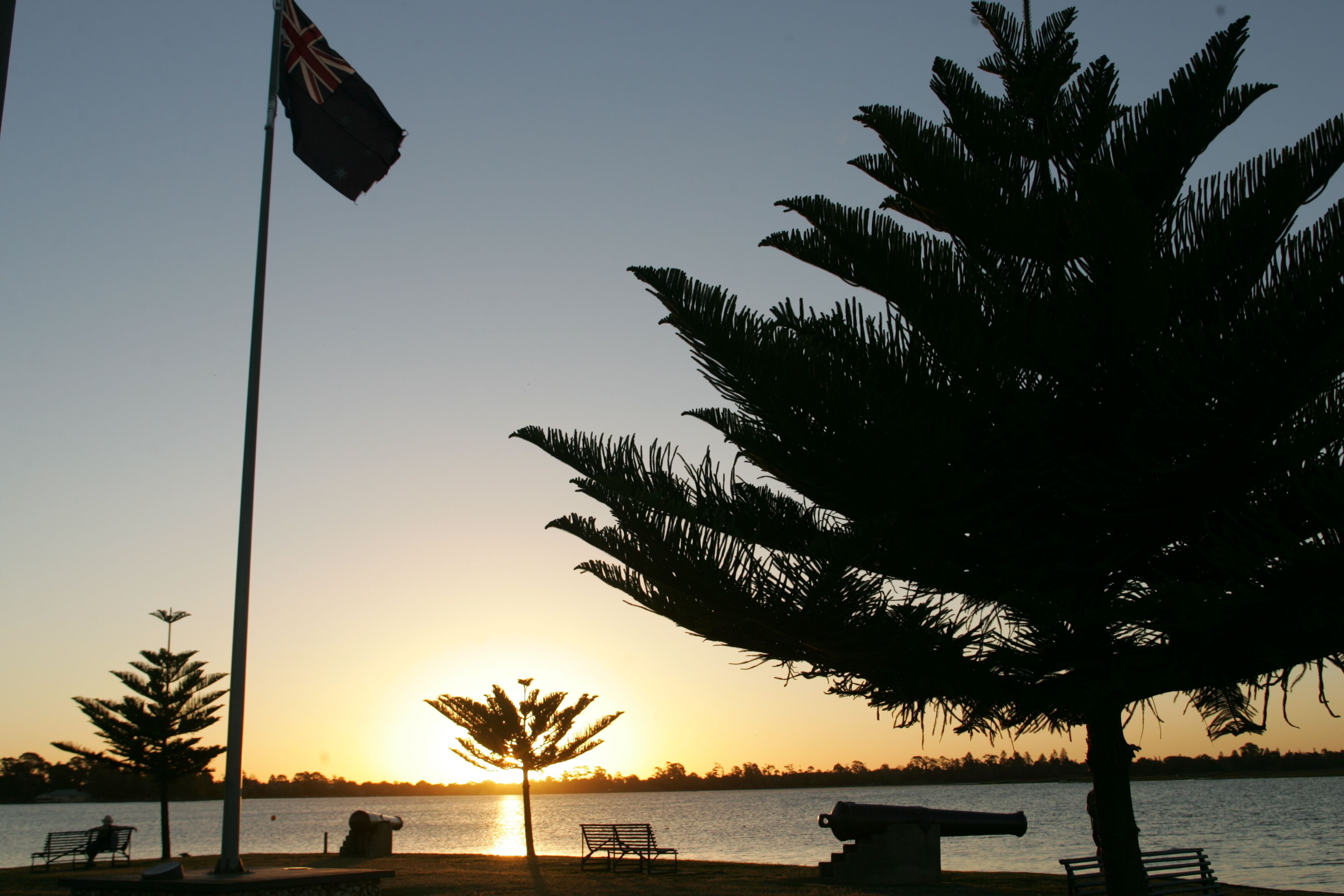 A sunset view with a flag, benches, and a palm tree by a body of water.