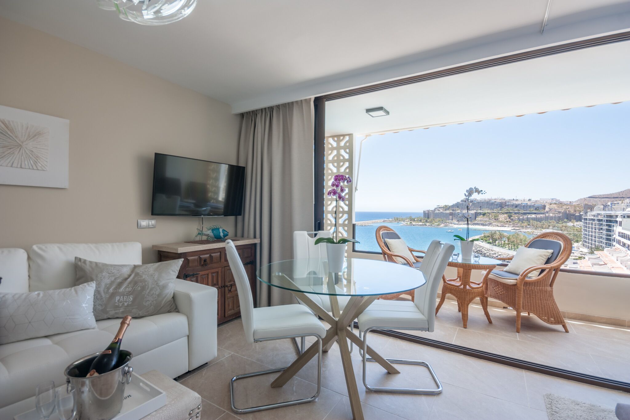 A modern living room with a glass table, white sofas, and a TV. A balcony with wicker chairs and a view of the sea is visible.
